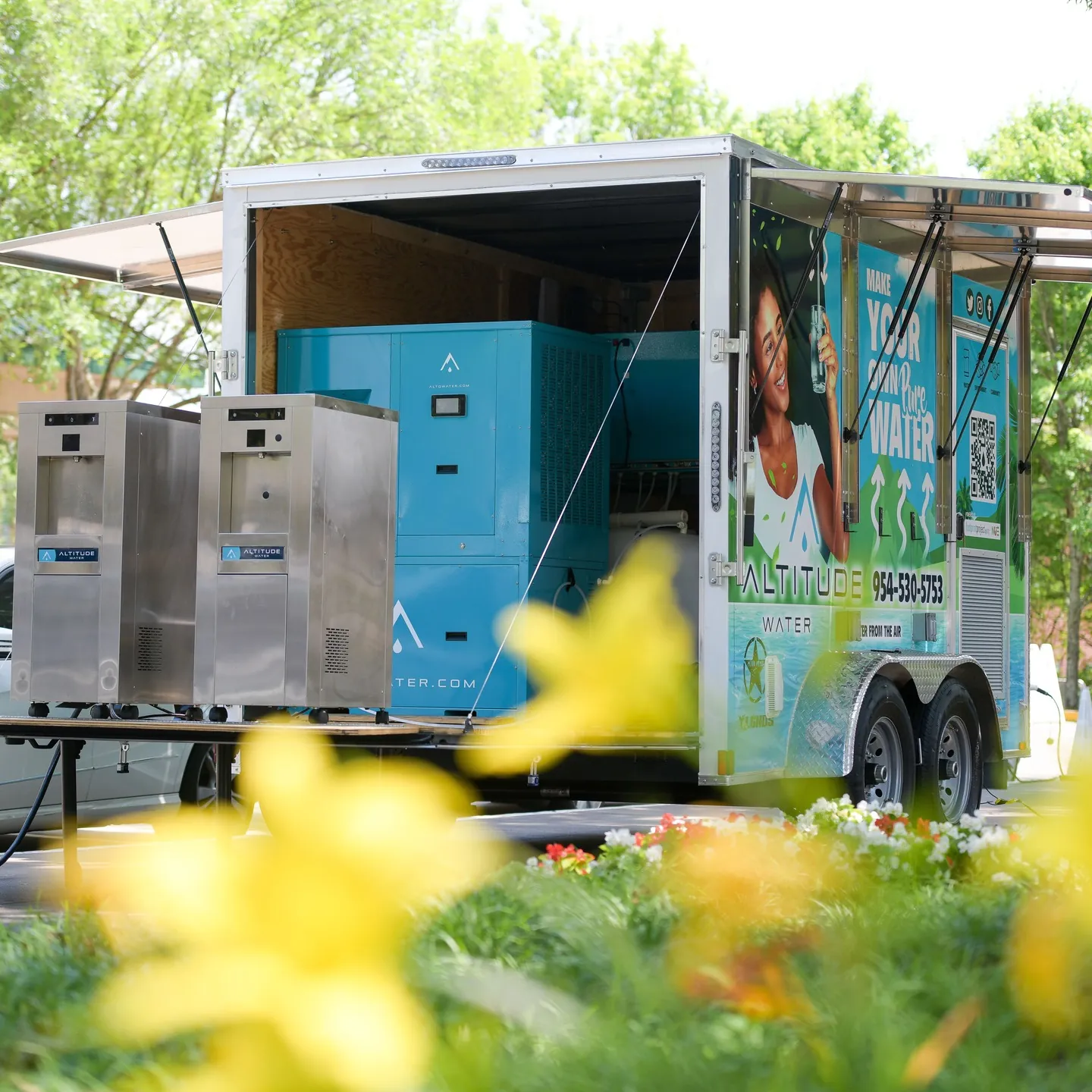 Open Altitude Water trailer with two stainless steel T-12 units in front and a blue T-series machine inside, yellow flowers blurred in the foreground.
