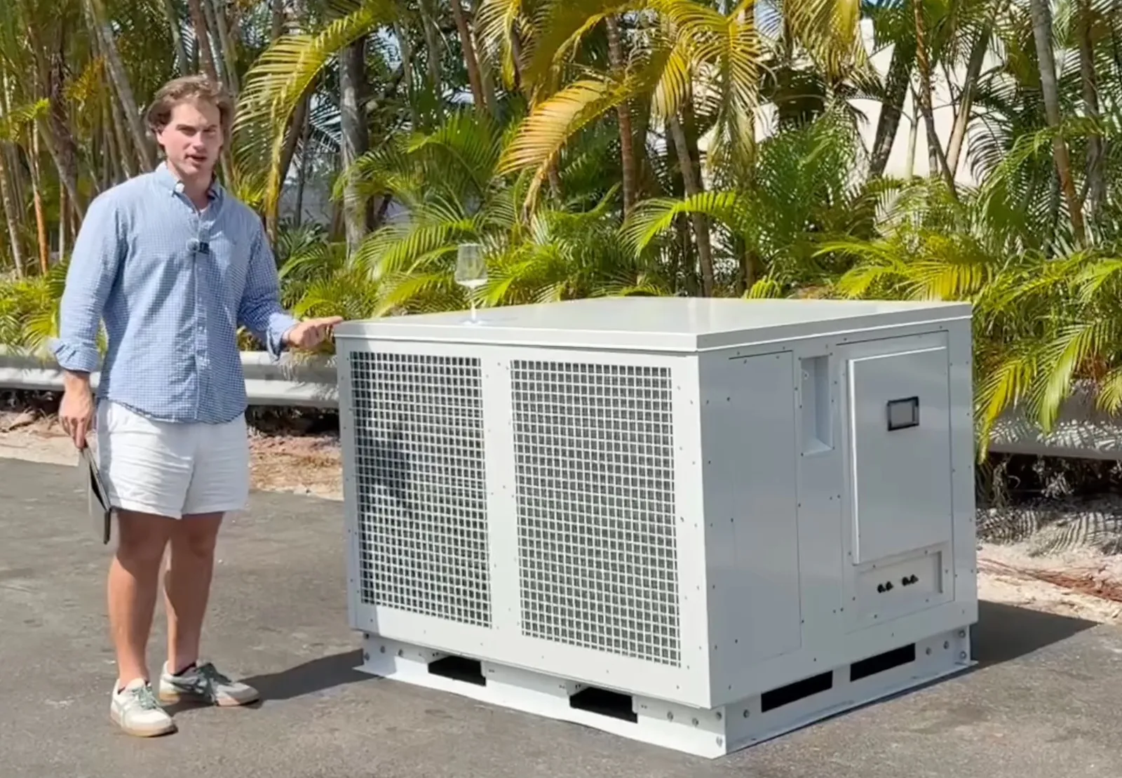Man in a blue shirt presenting a white Altitude Water commercial generator, with a glass of water on top, among palm trees.