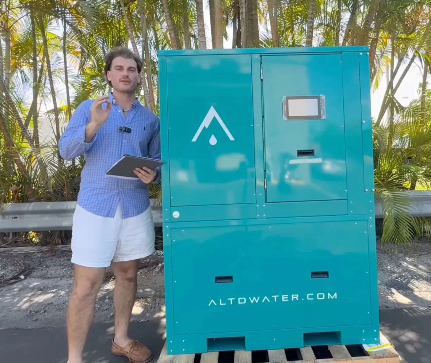 Man holding a tablet next to a teal Altitude Water commercial generator on a pallet, palm trees behind.
