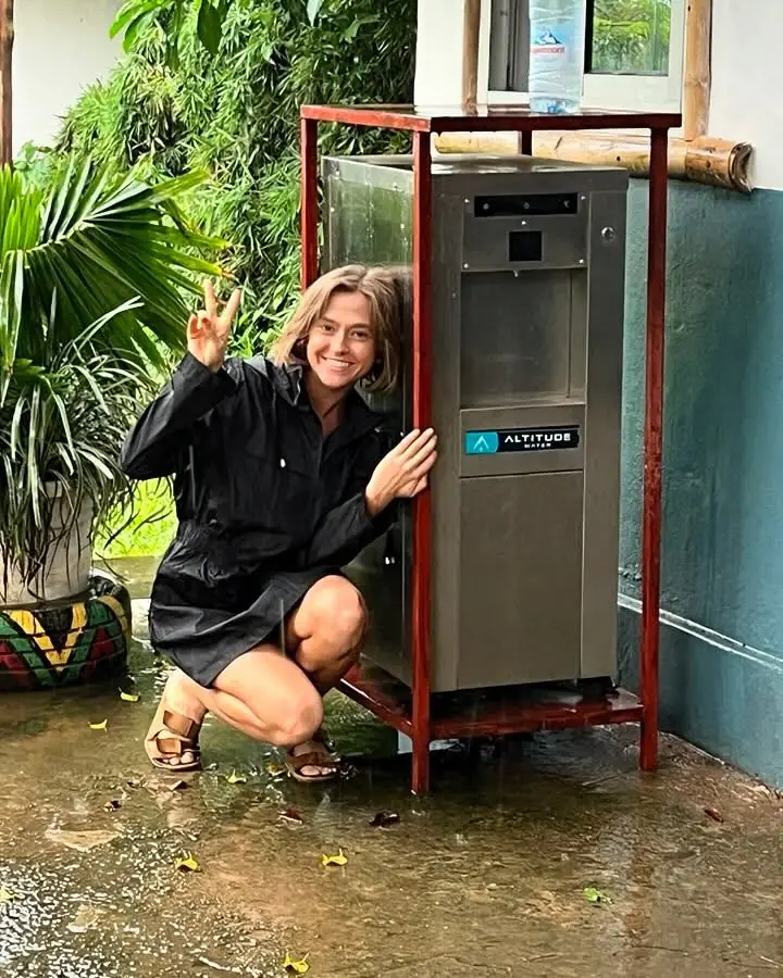 Smiling woman in a raincoat crouching next to a T-12 on a red metal stand, on a patio wet from the rain.